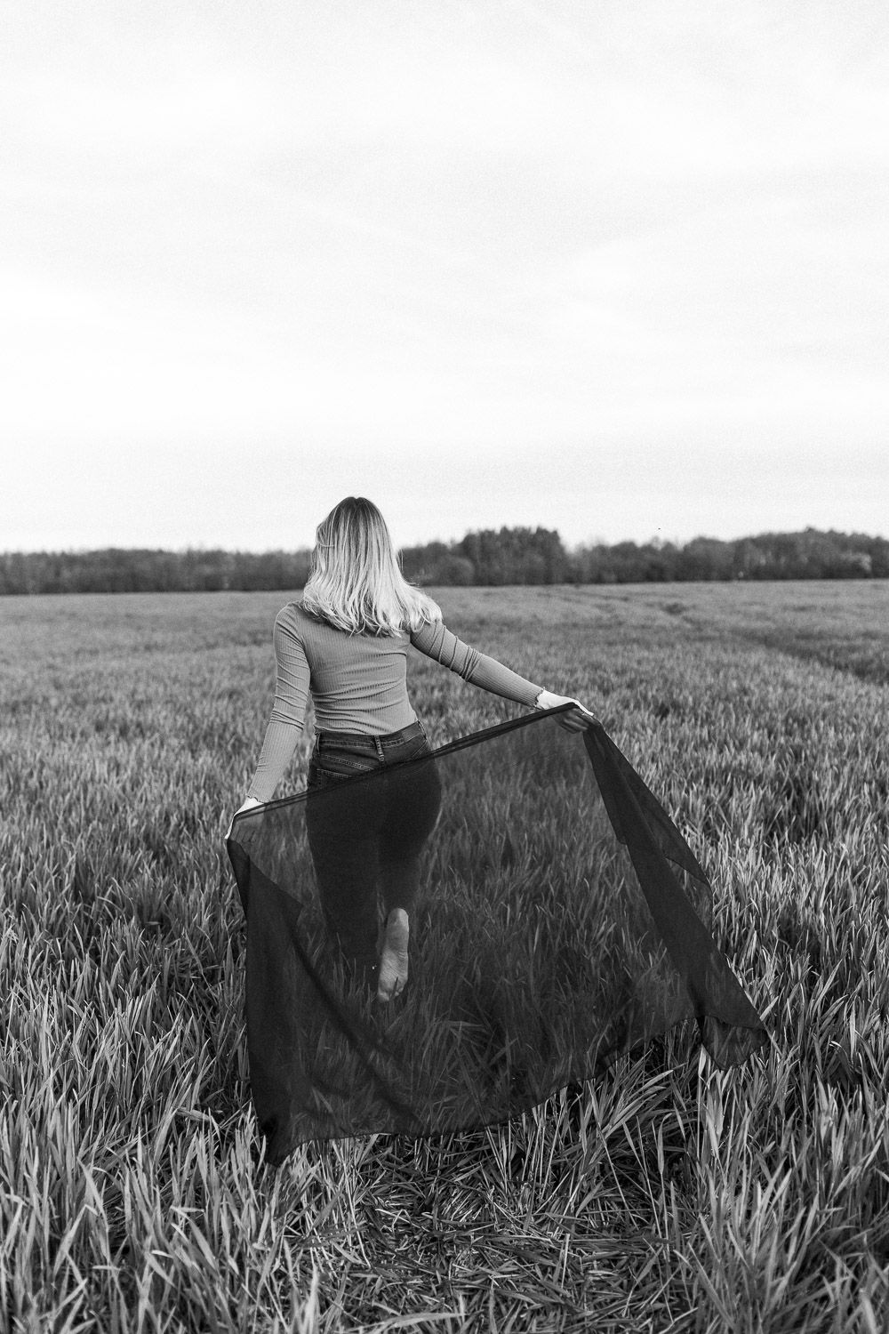 Figure stands in field holding flowing black fabric while facing away from camera.