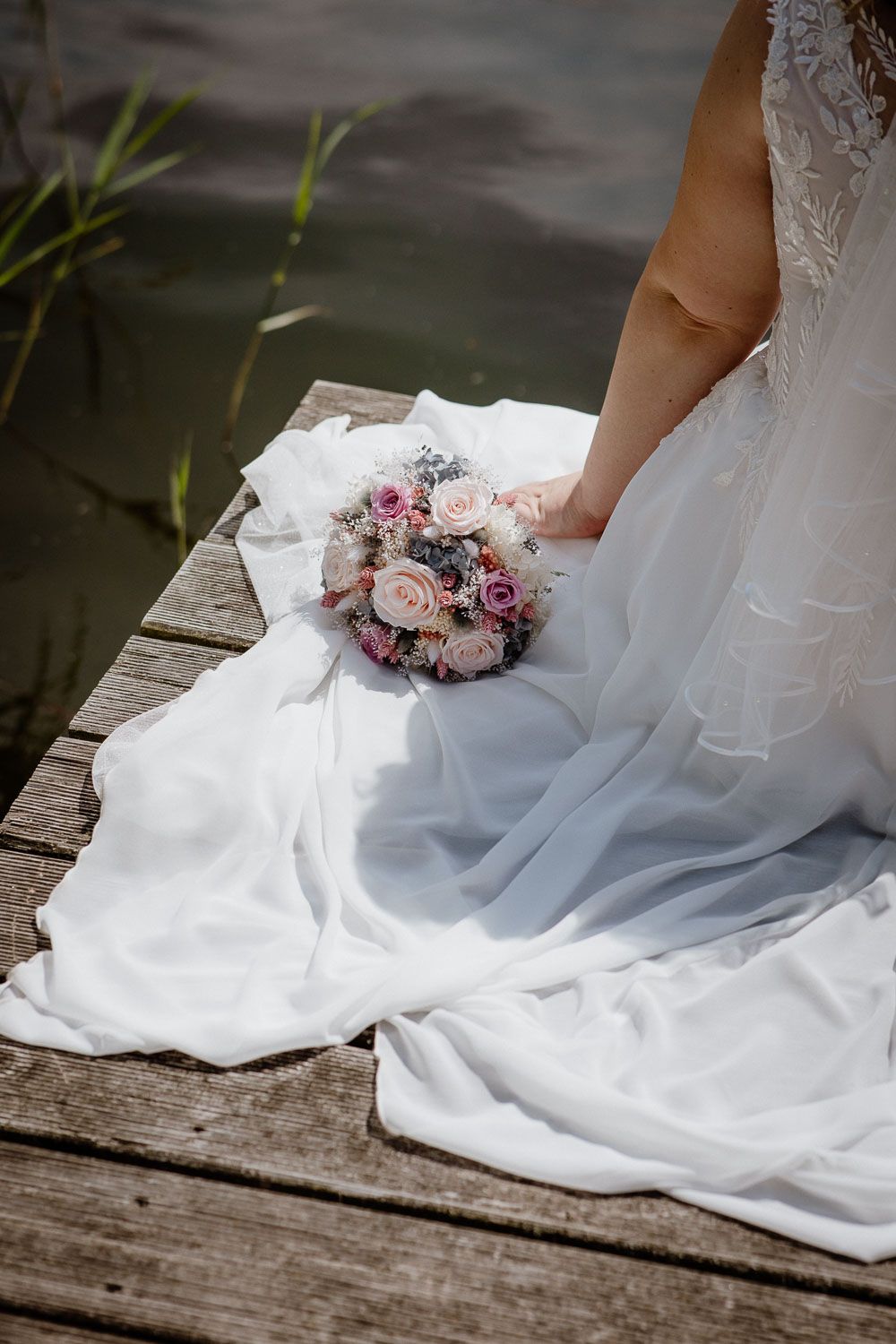 A white wedding dress and pastel bouquet rest on a wooden dock by the water.