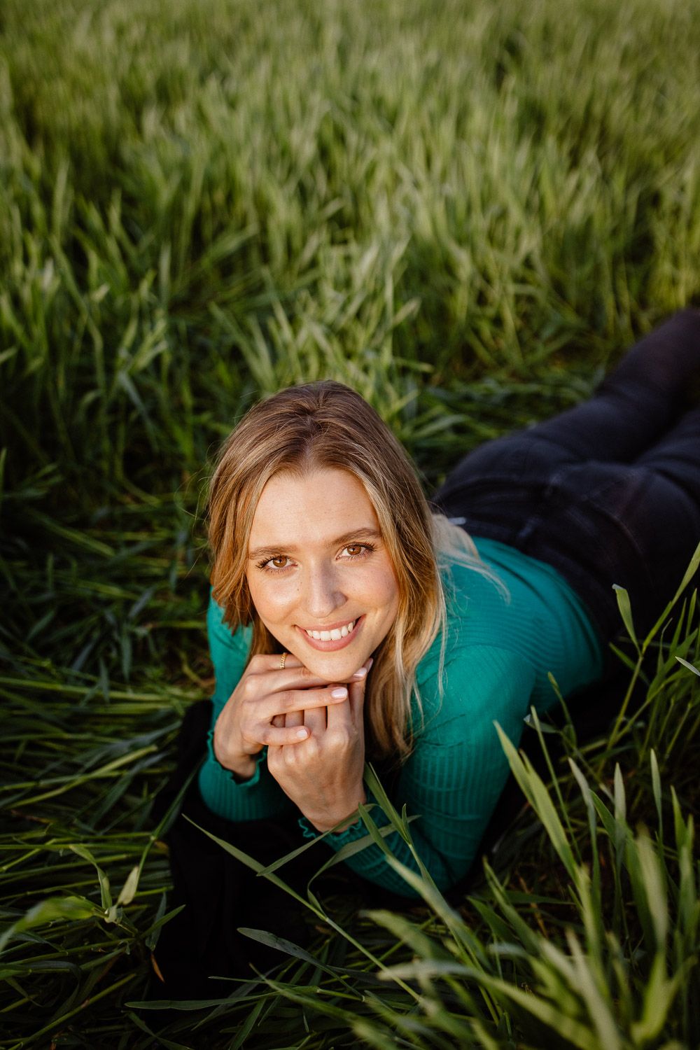 Relaxed portrait laying in tall green grass wearing turquoise sweater with natural lighting.