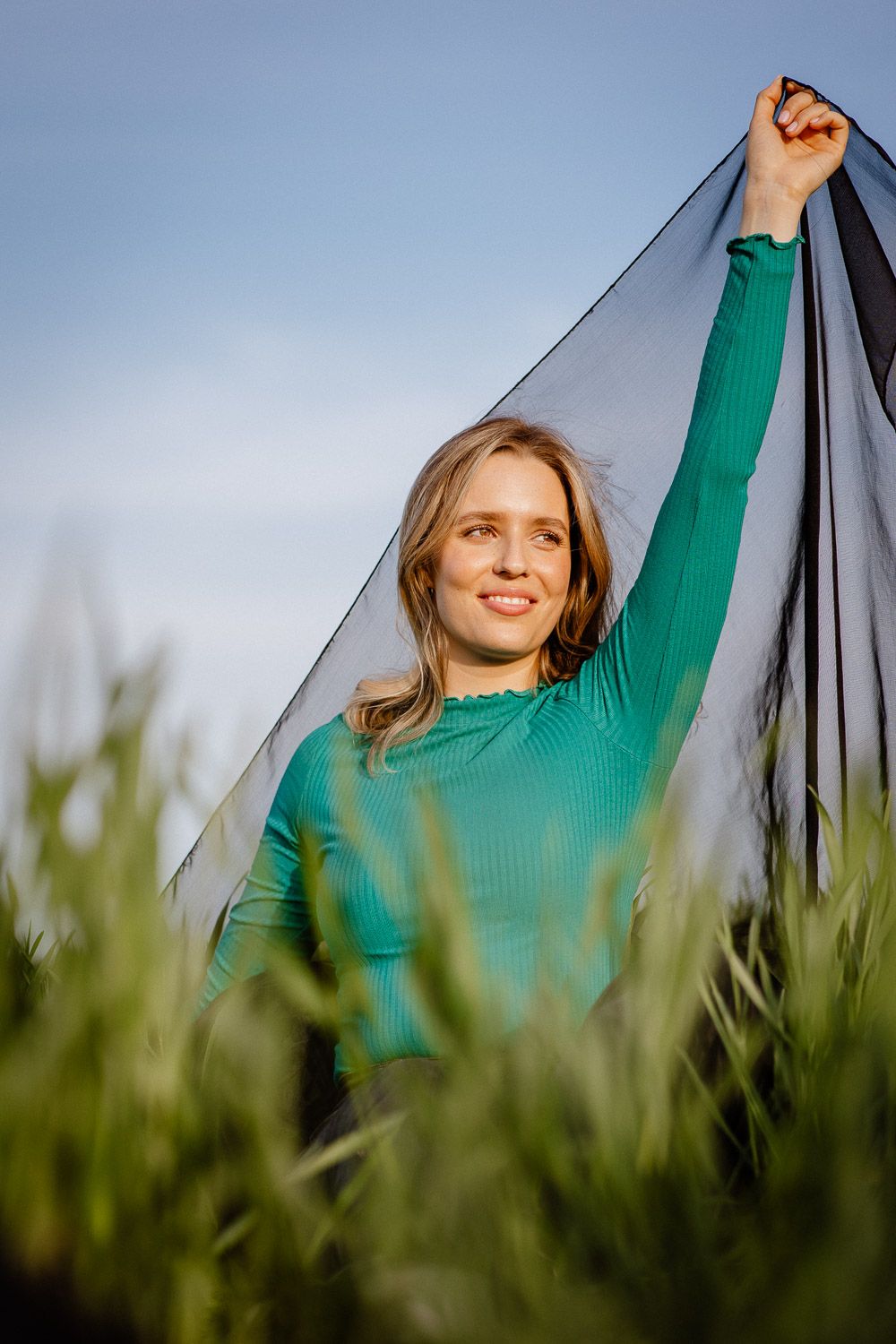 A figure in a green sweater holds up a dark fabric against a blue sky in a grassy field.