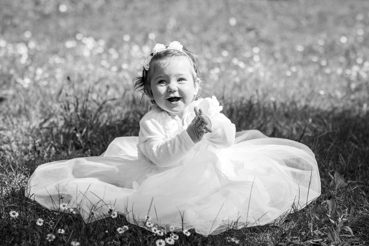 Black and white portrait of baby in white dress sitting among flowers.