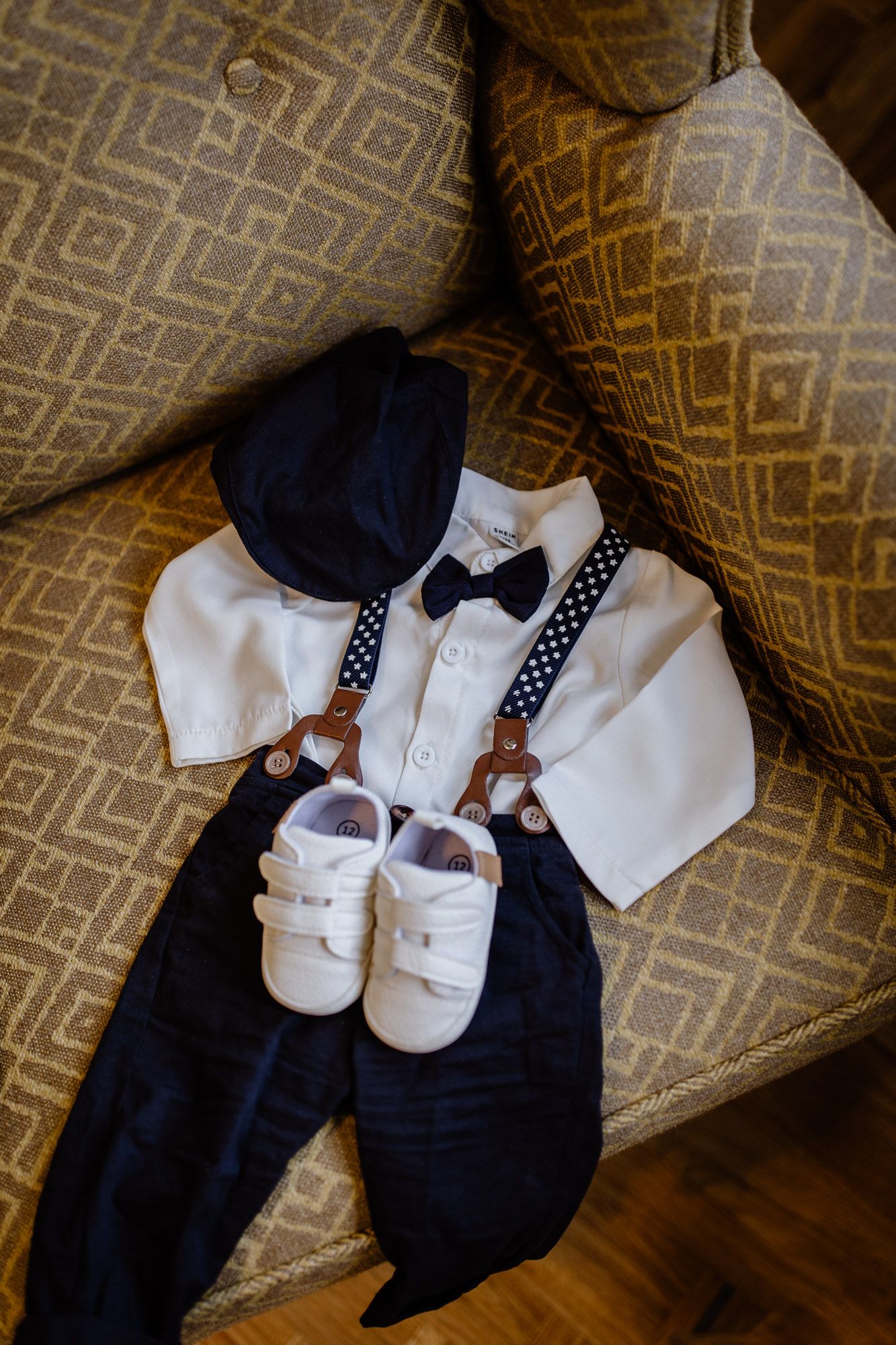 Baby formal wear including white shoes, navy suit, and polka dot bow tie arranged on patterned upholstery.