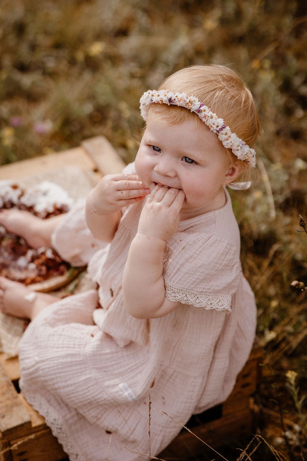A child in a white lace dress and floral headband sits outdoors with messy hands and a playful expression.