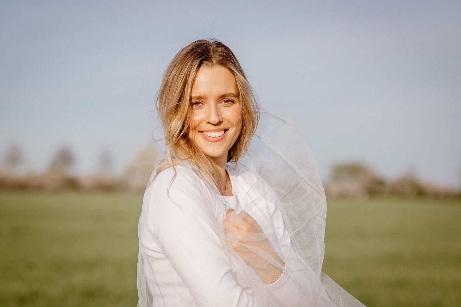 A person in a white dress stands in a meadow during golden hour while a light veil flows in the breeze.