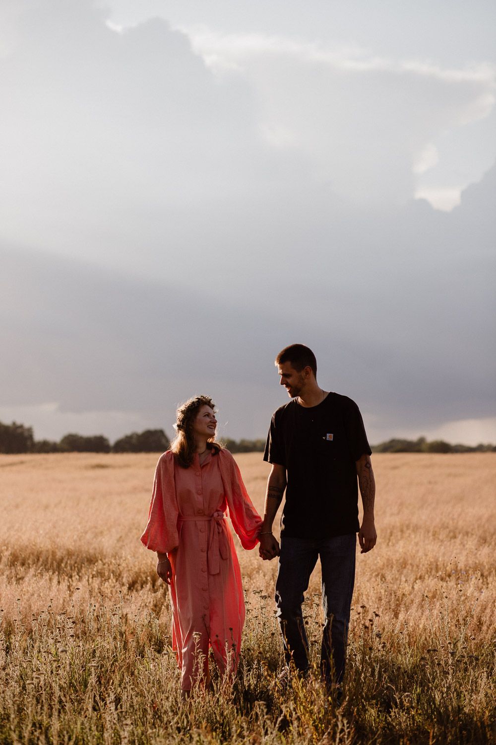 Couple walking through a golden wheat field at sunset against cloudy skies.