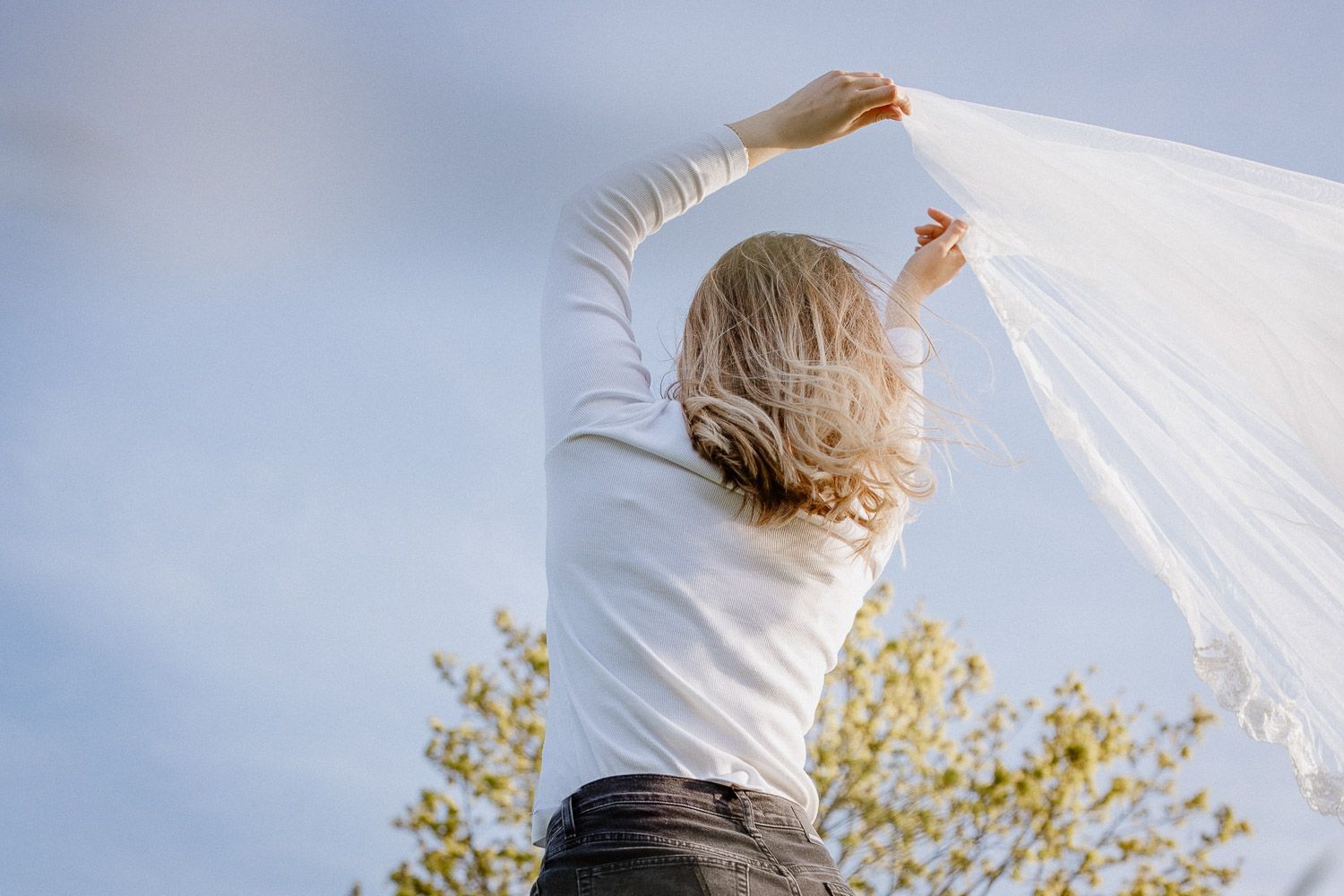 Figure raises white fabric overhead against blue sky with trees in background.