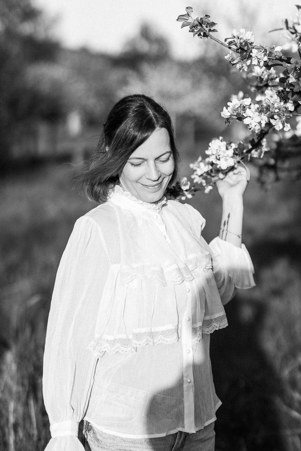 A romantic black and white portrait of someone in a white lace blouse standing by spring blossoms in nature.