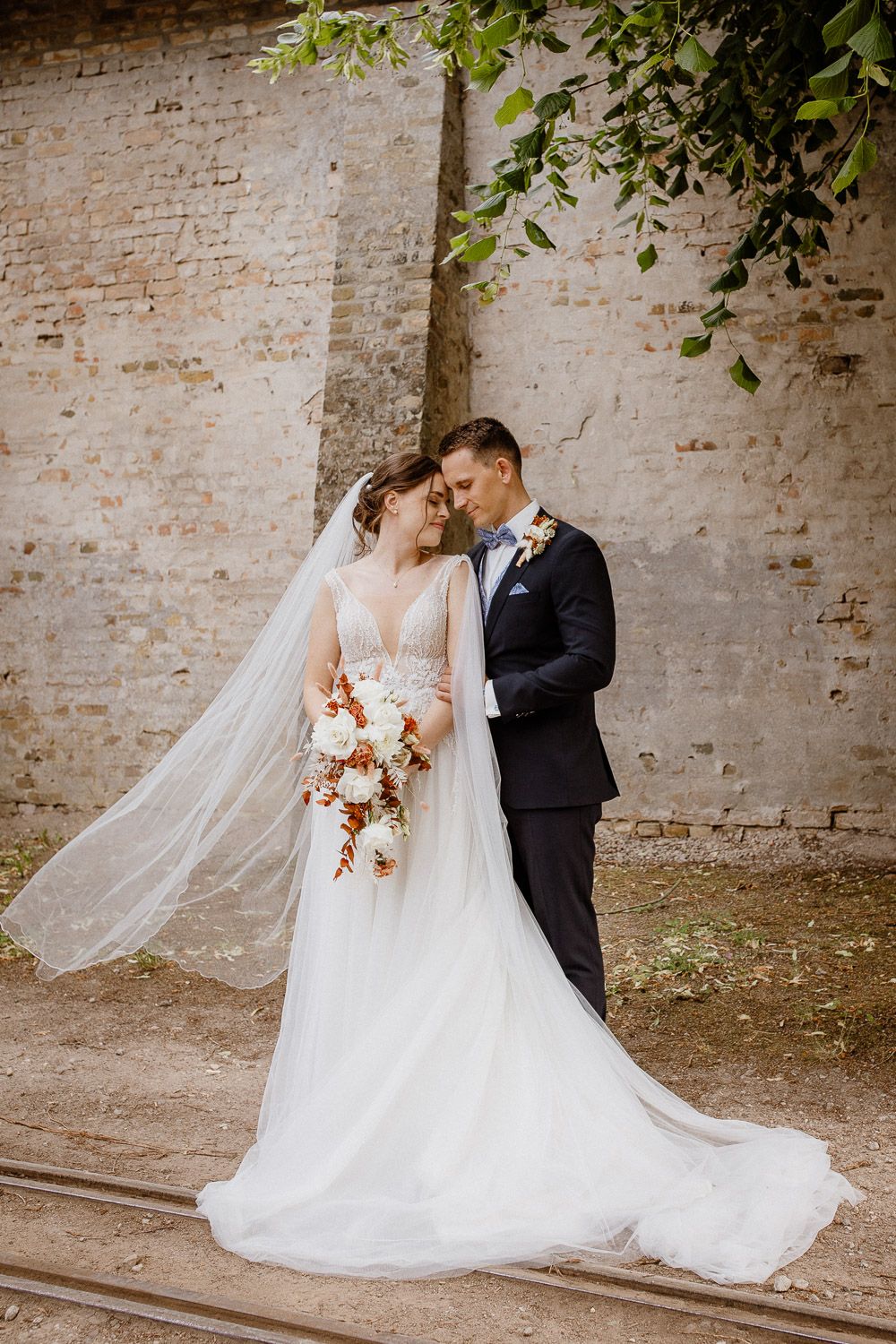 Wedding couple embracing against rustic brick wall with flowing veil and floral bouquet.