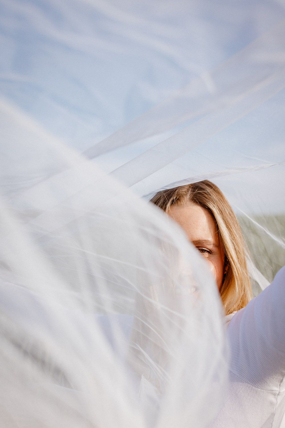 Close-up portrait partially obscured by flowing white veil against blue sky.