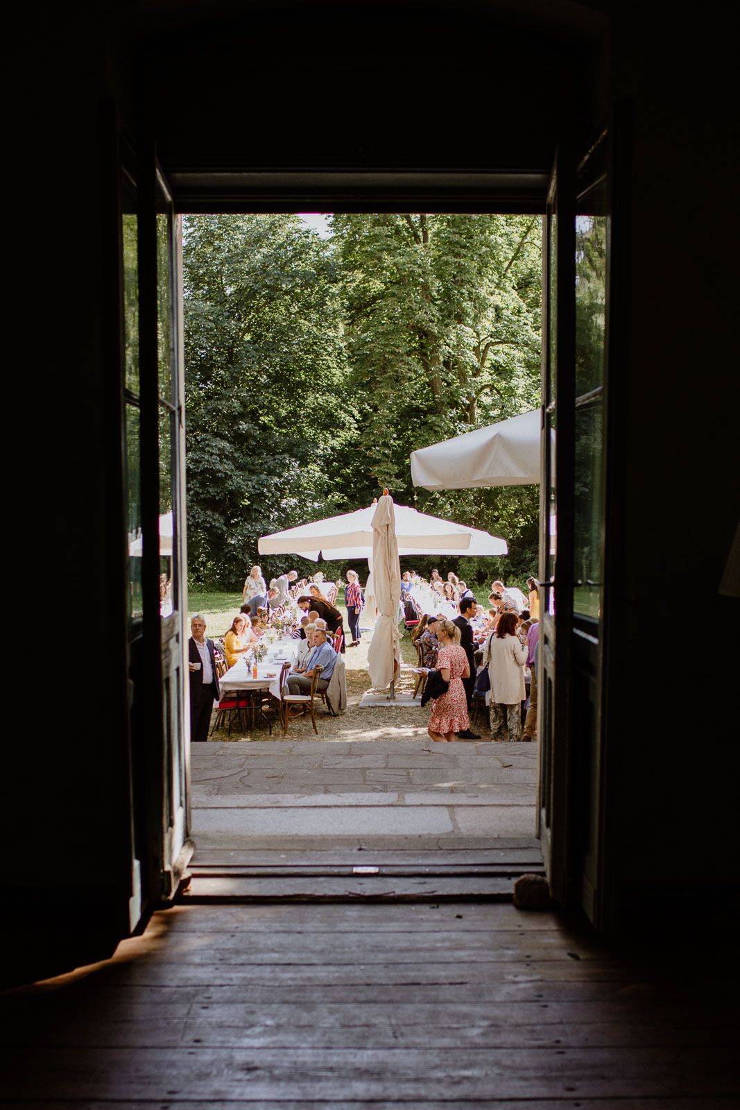 View through dark doorway of outdoor wedding reception with white umbrellas and guests gathered in garden setting.
