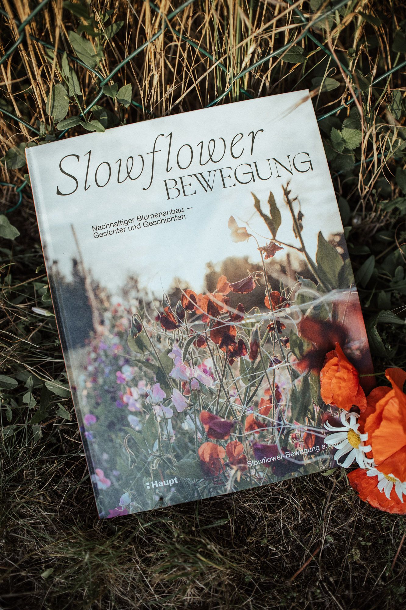 A book titled 'Slowflower Bewegung' displayed on grass with dried flowers and orange petals in the background.