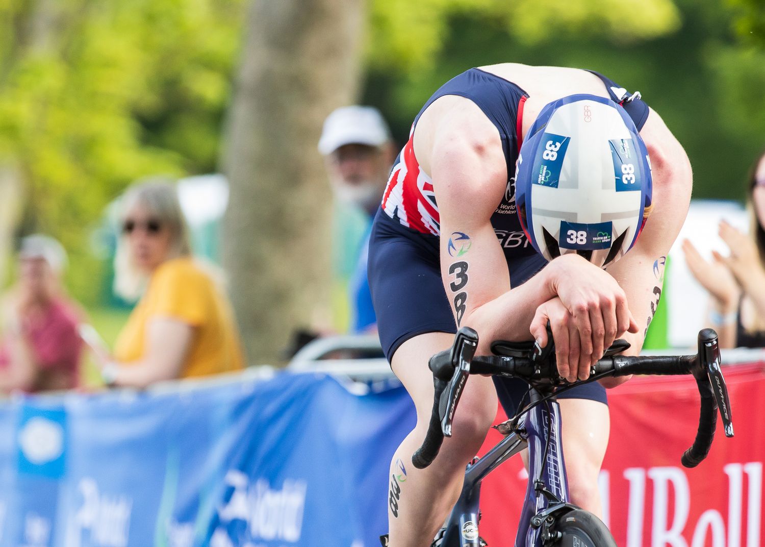 Cyclist in aerodynamic position on racing bike during competitive triathlon event with spectators in background.