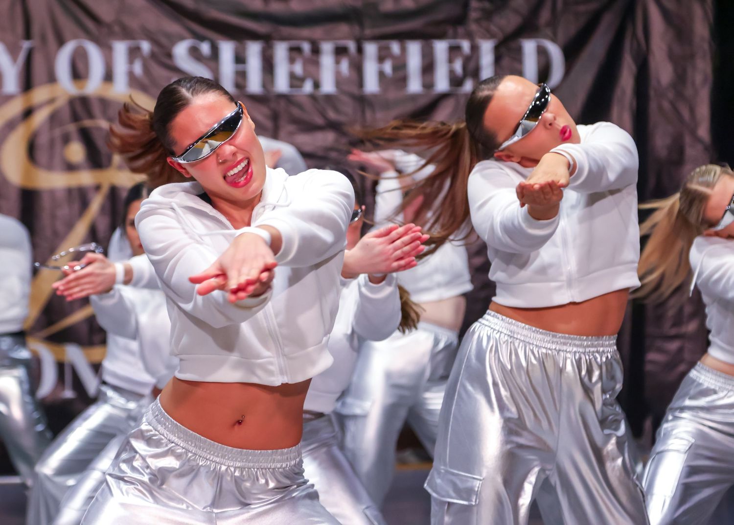 Dancers in white tops perform an energetic routine with animated expressions on stage.