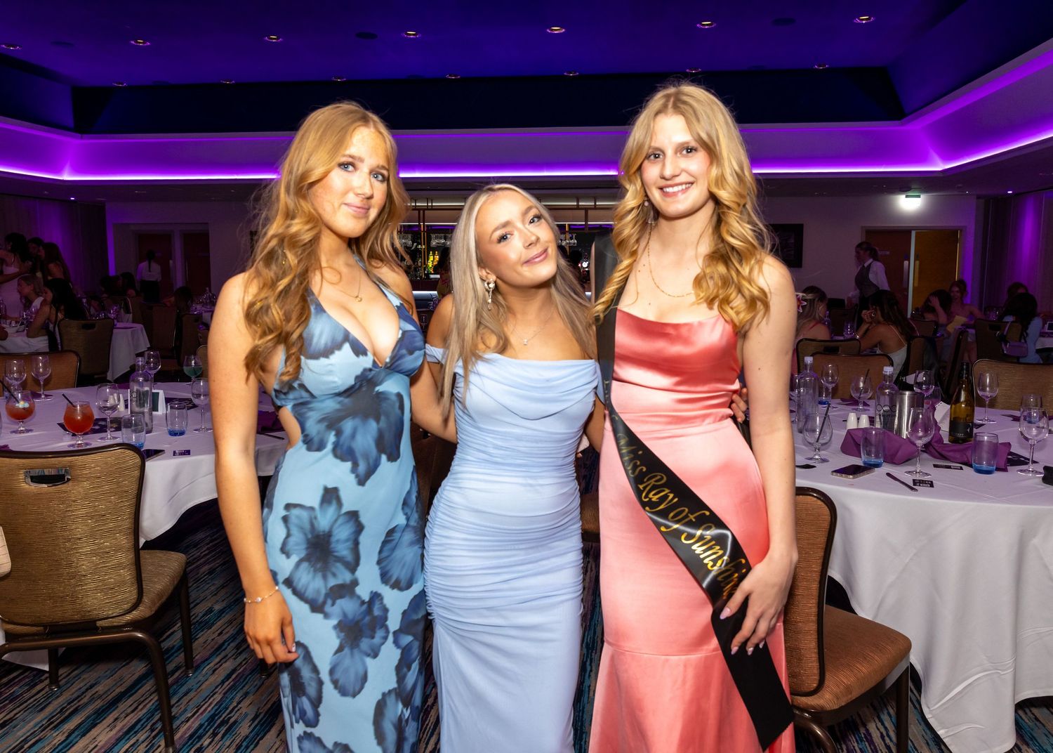 Three friends in elegant formal dresses pose together at an upscale event with purple lighting.