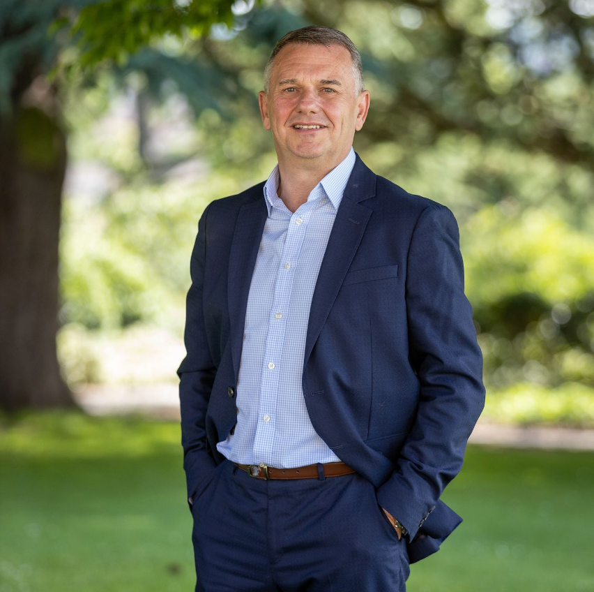 Professional wearing navy suit and light blue shirt stands outdoors in park setting.