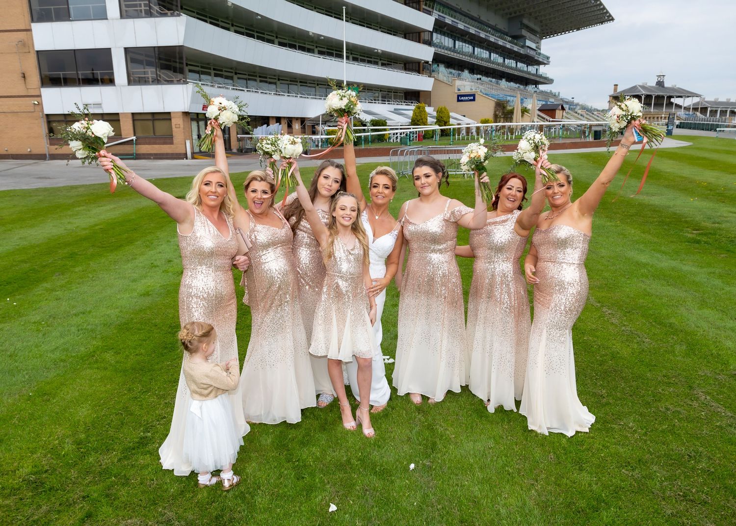 Bridesmaids in light colored dresses celebrating on green lawn at racecourse venue.