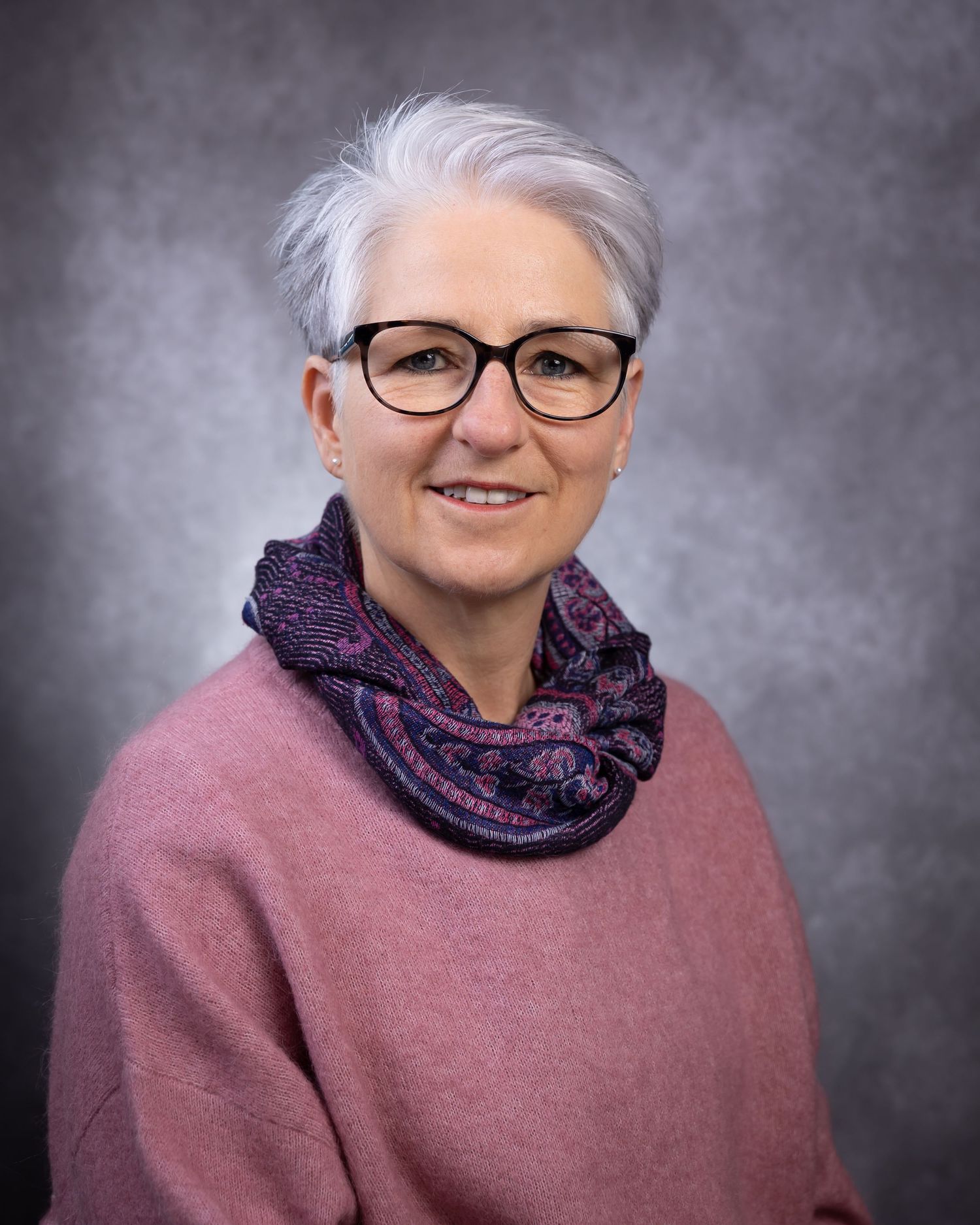Professional studio portrait of someone wearing a pink sweater and patterned scarf against a gray backdrop.