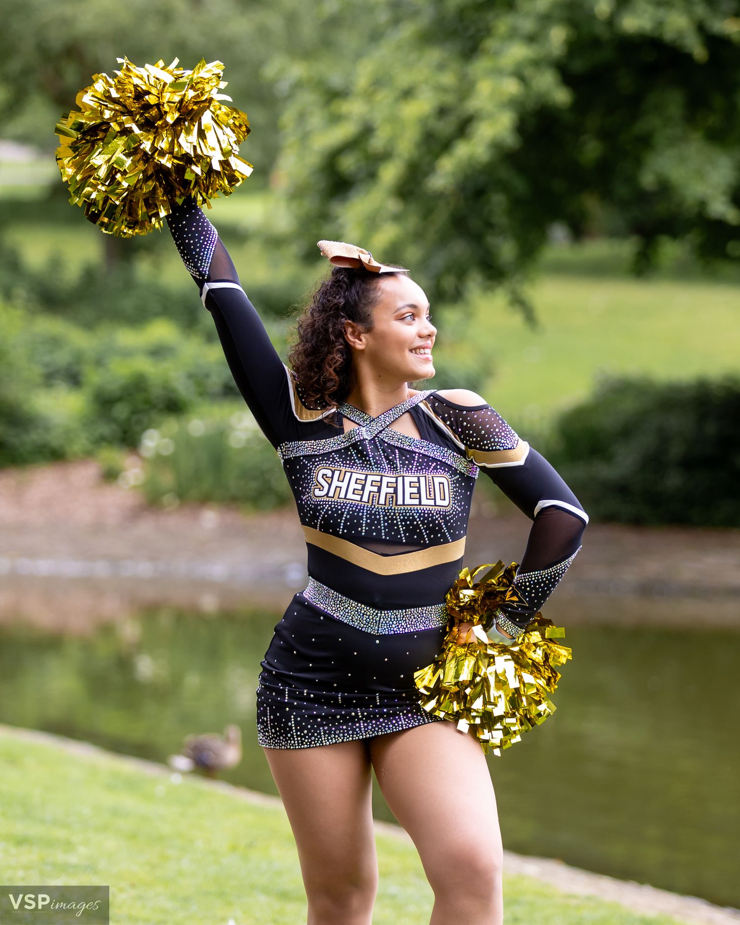 Cheerleader in black and gold uniform performing with pom poms by a lake.
