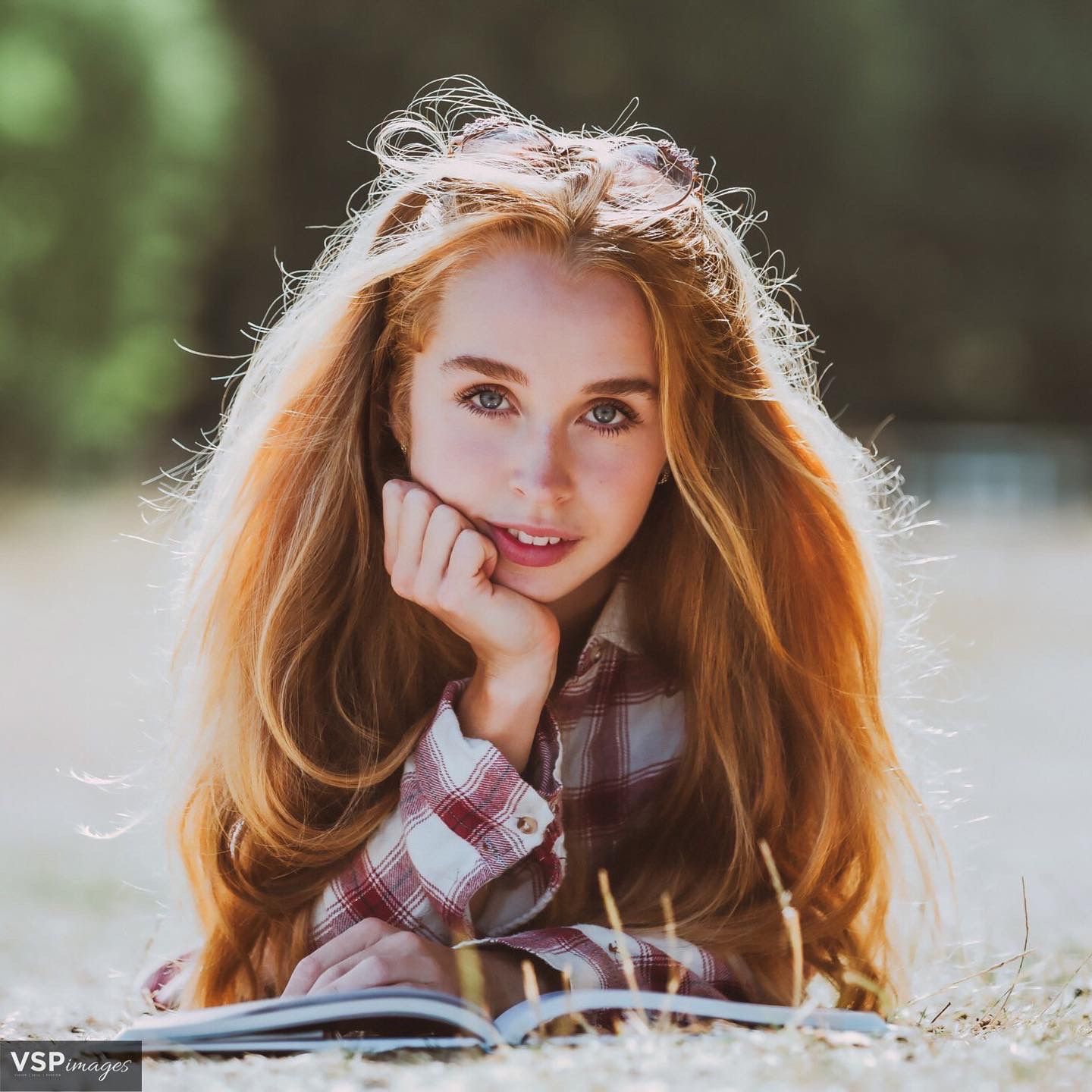 Young redhead in plaid shirt lies on ground reading while resting chin on hand with soft sunlight behind.