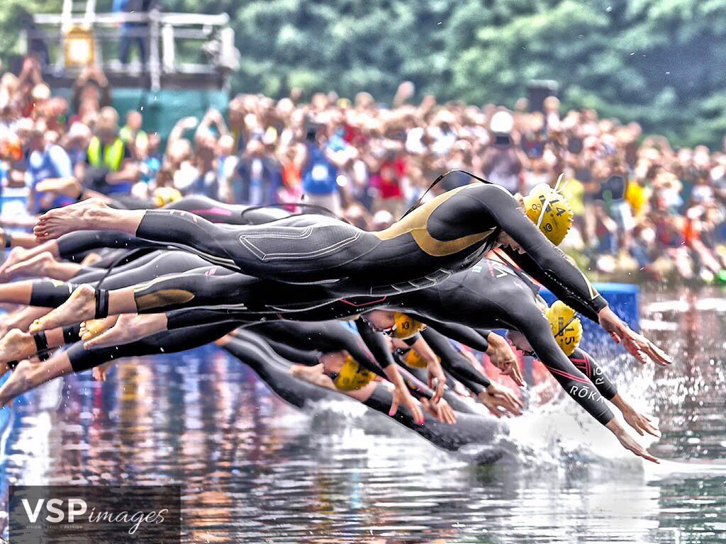 Athletes dive into the water at the start of a competitive TRIATHLON race on a sunny day at an outdoor pool.