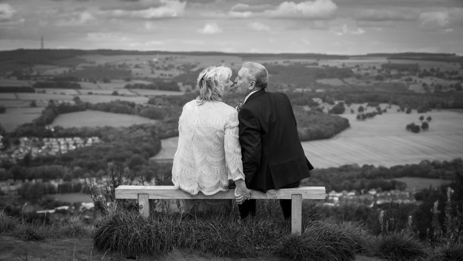 A couple sits together on a bench overlooking a scenic countryside valley view.