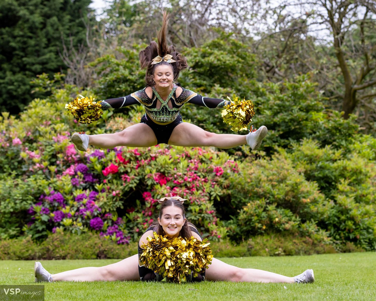 Two cheerleaders performing synchronized splits pose among colorful garden flowers.
