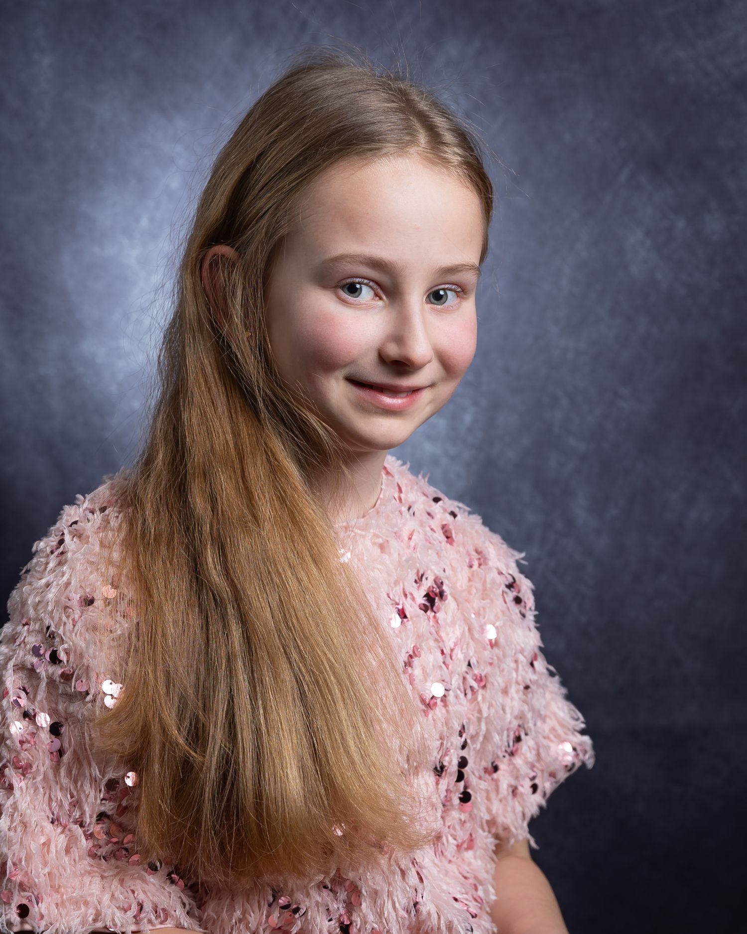 Studio portrait of a young person with long hair wearing a pink lace dress against a dark textured background.
