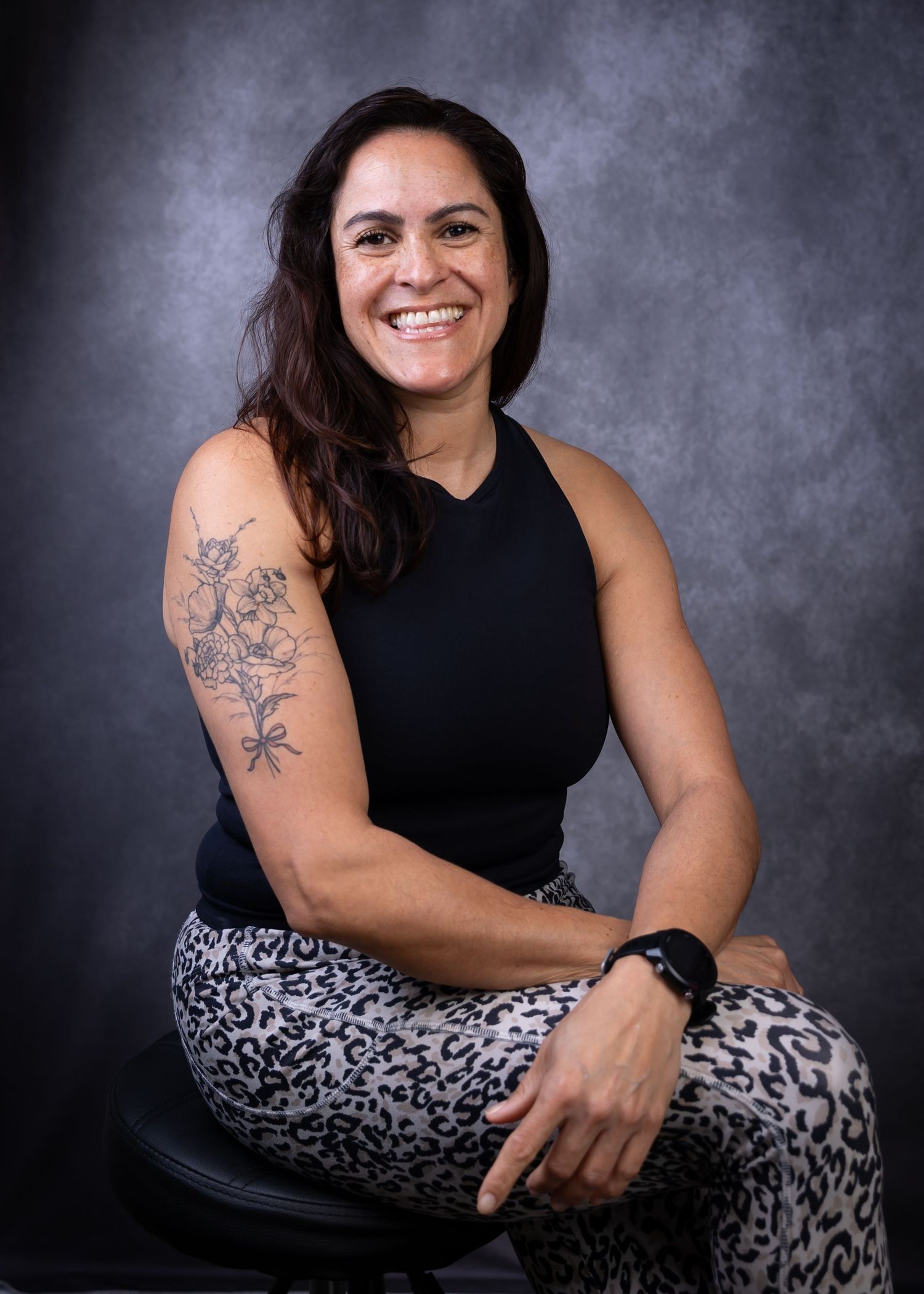 Professional studio portrait of someone in a black sleeveless top and leopard print pants showing a floral arm tattoo.