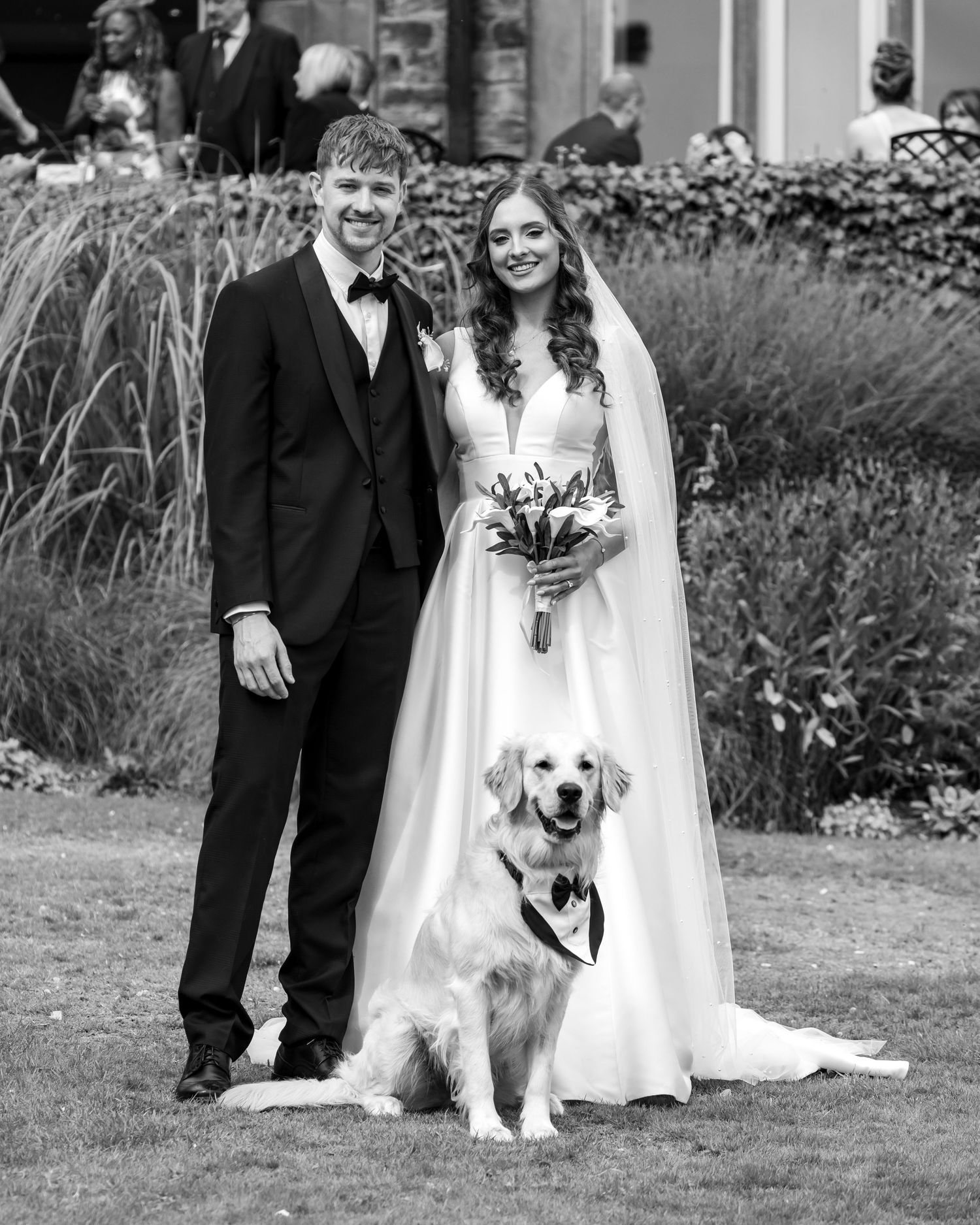 Couple in wedding attire poses with their golden retriever dog in outdoor garden setting for wedding portraits.