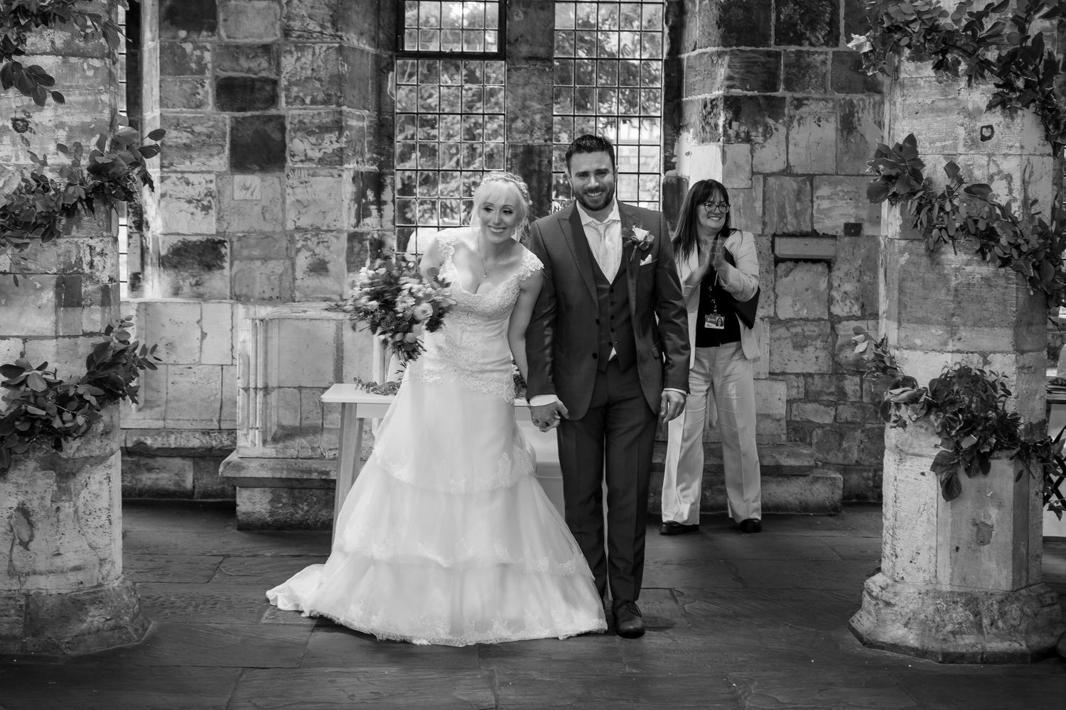 Black and white photo of a joyful couple walking together through an elegant stone archway on their wedding day.
