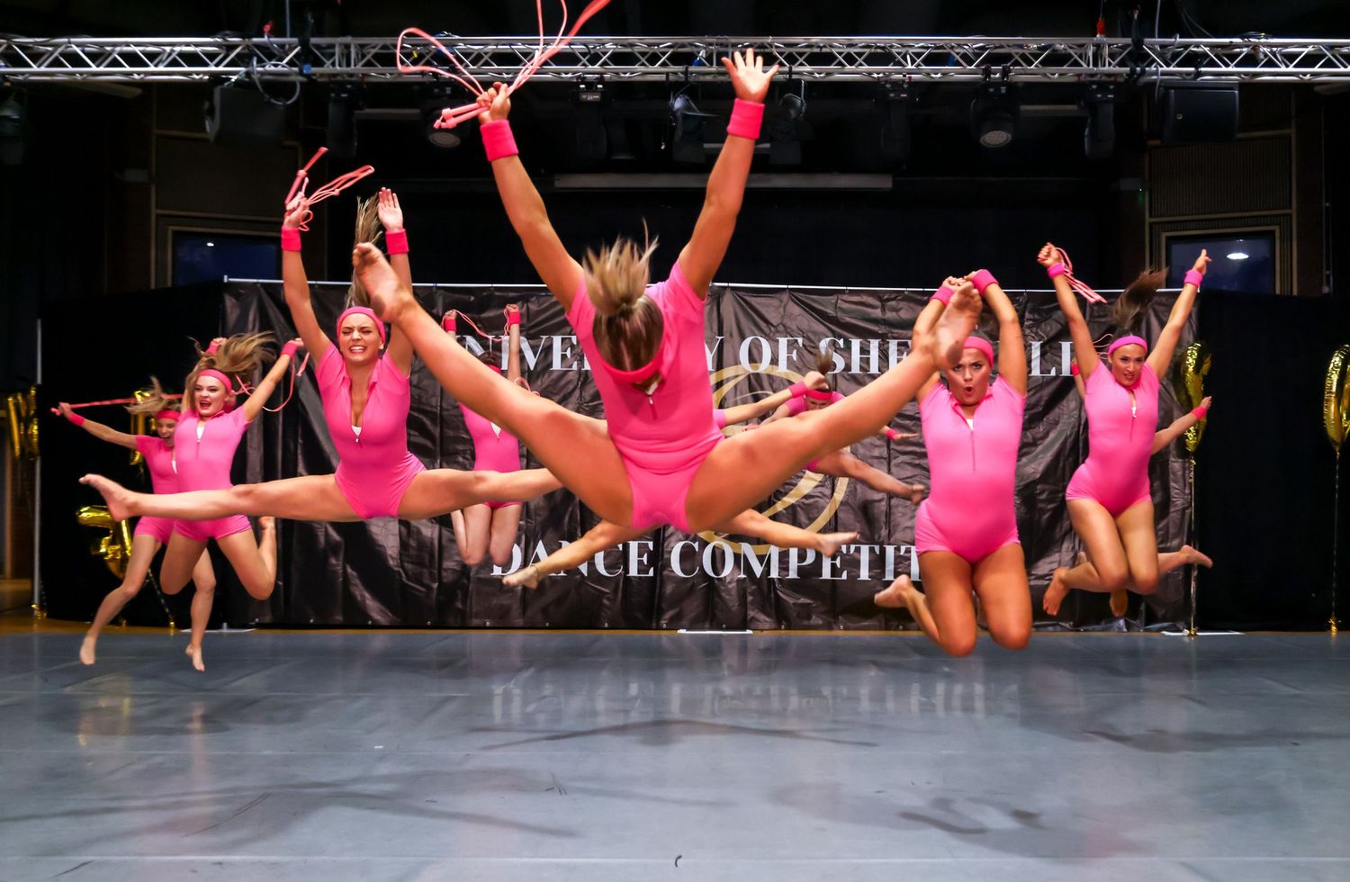Energetic dancers in pink costumes leap simultaneously during a stage performance.