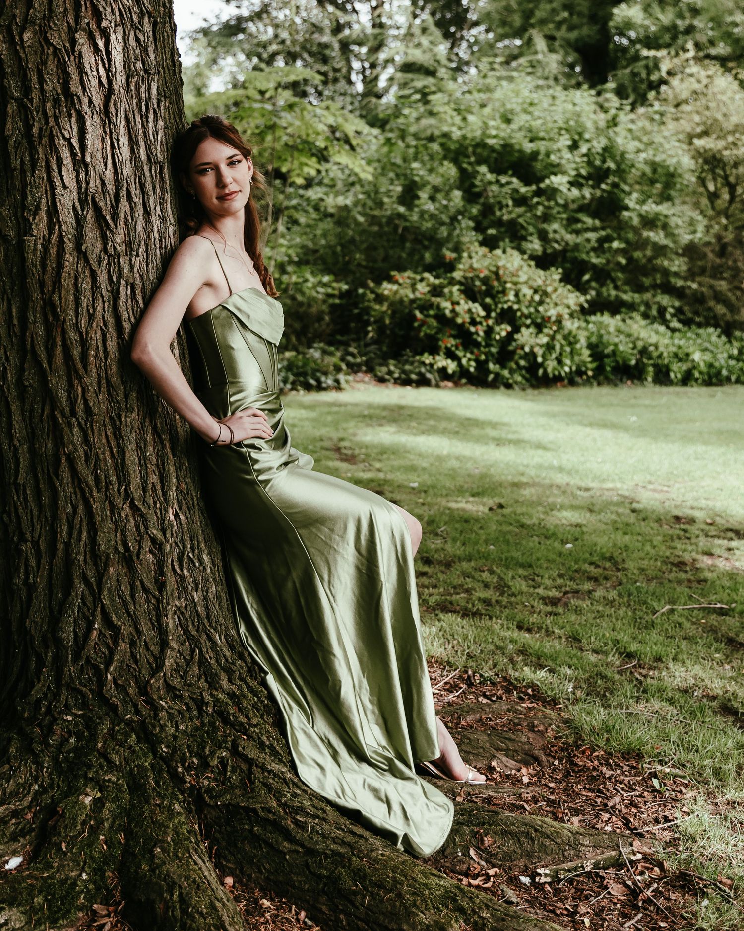A woman in a sage green dress sits elegantly against a large tree trunk in a serene garden setting.
