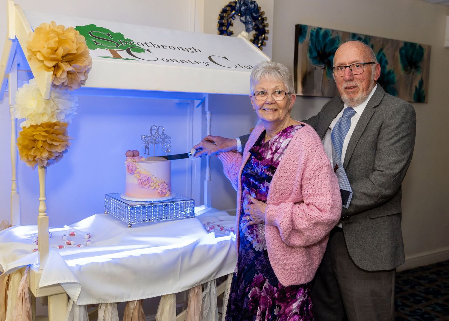 Elderly couple cutting a celebration cake at a Breakthrough Dementia Care event, decorated with flowers.