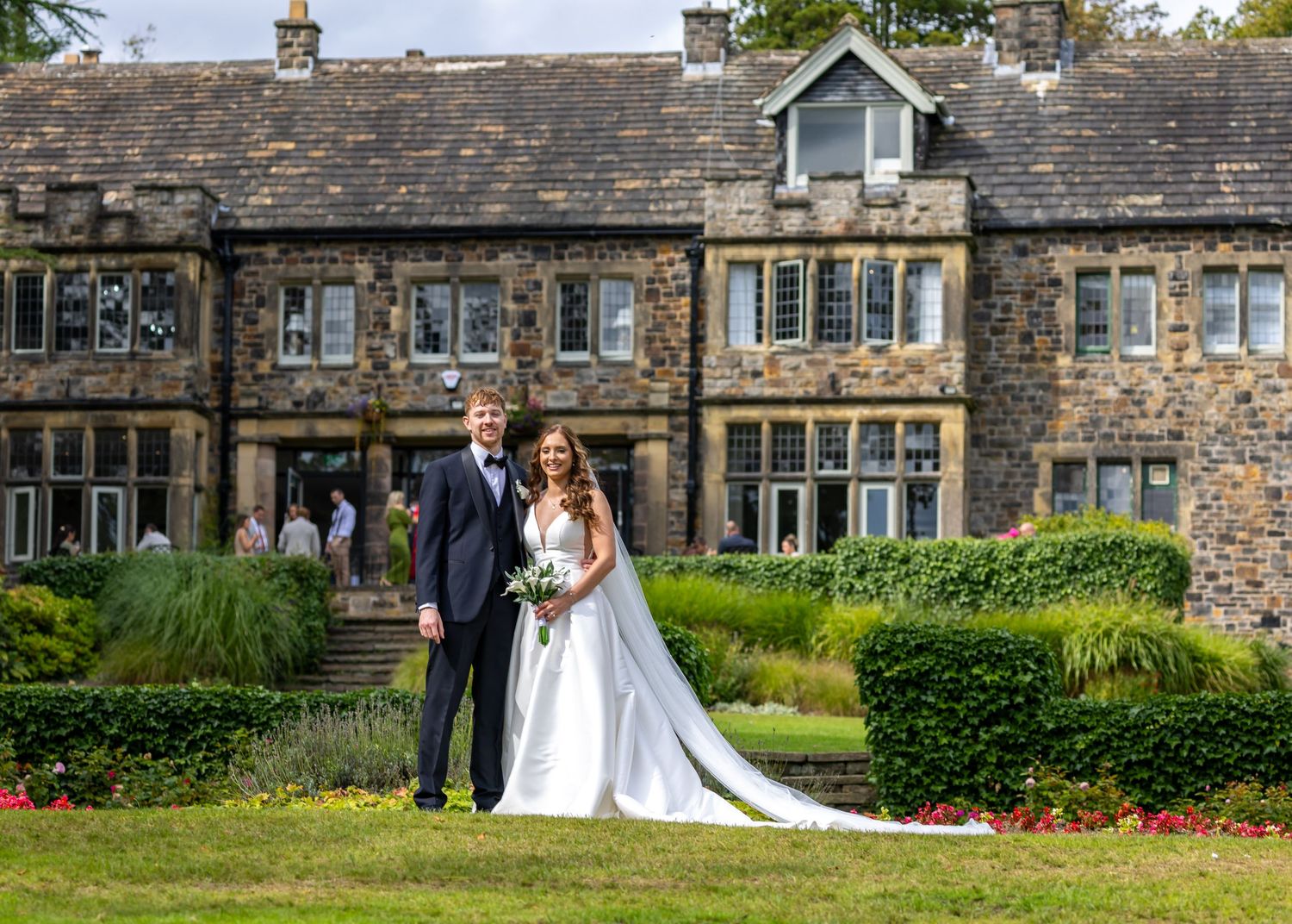 Bride and groom pose in wedding attire before a grand stone manor house surrounded by lush gardens.
