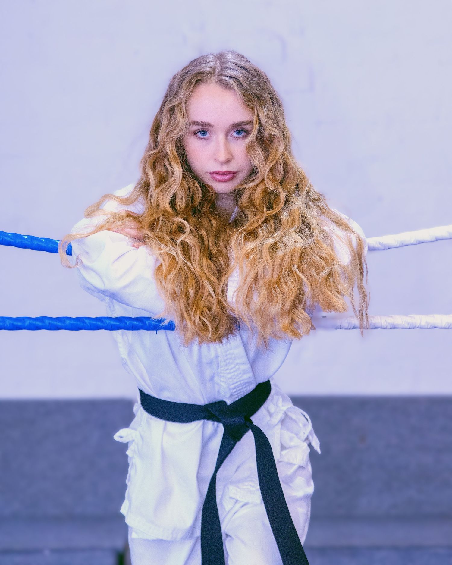 Martial artist in white uniform with black belt poses against blue ropes.