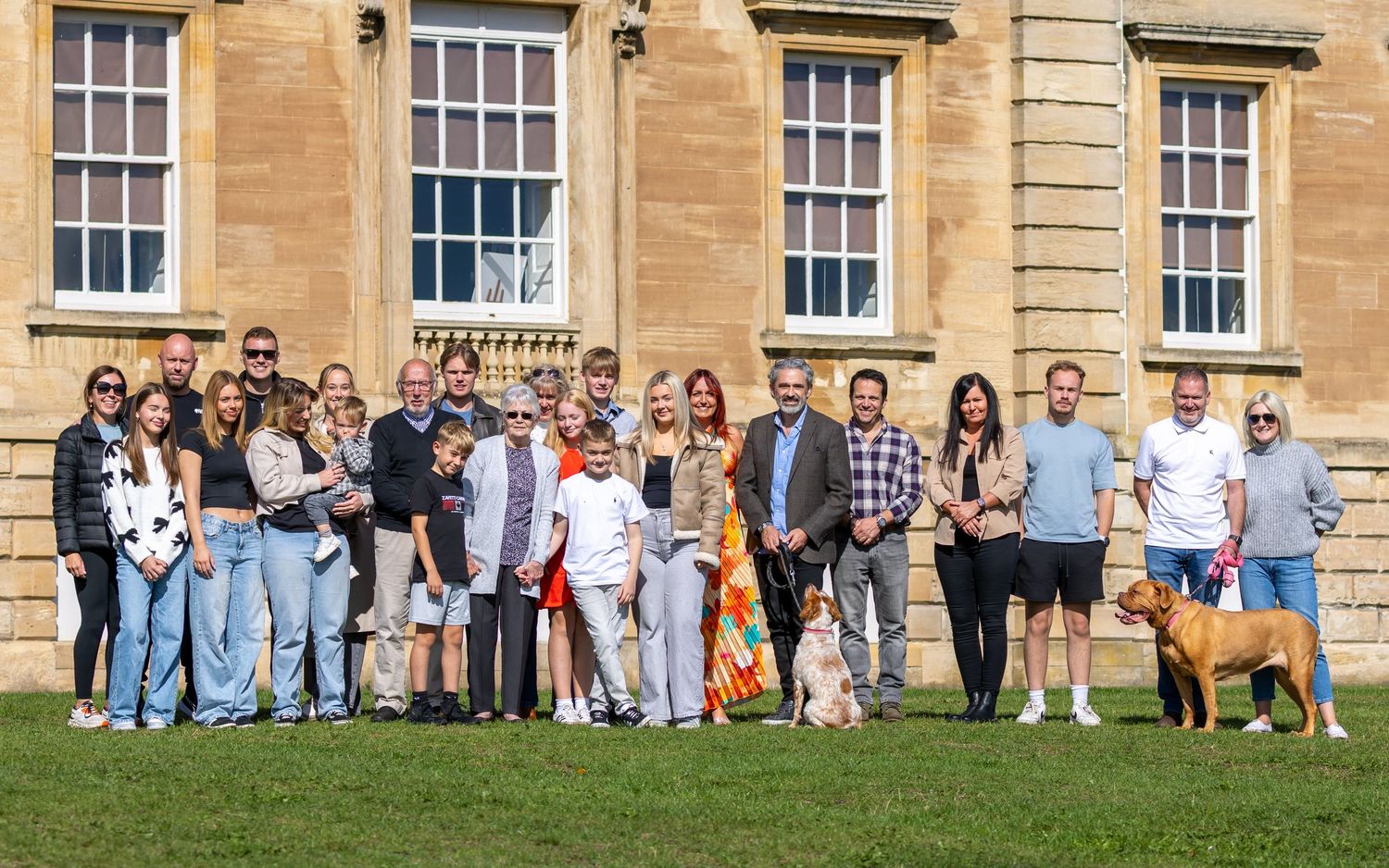 A group of people and dogs gather for a photo on a sunny day outside a historic stone building with large windows.
