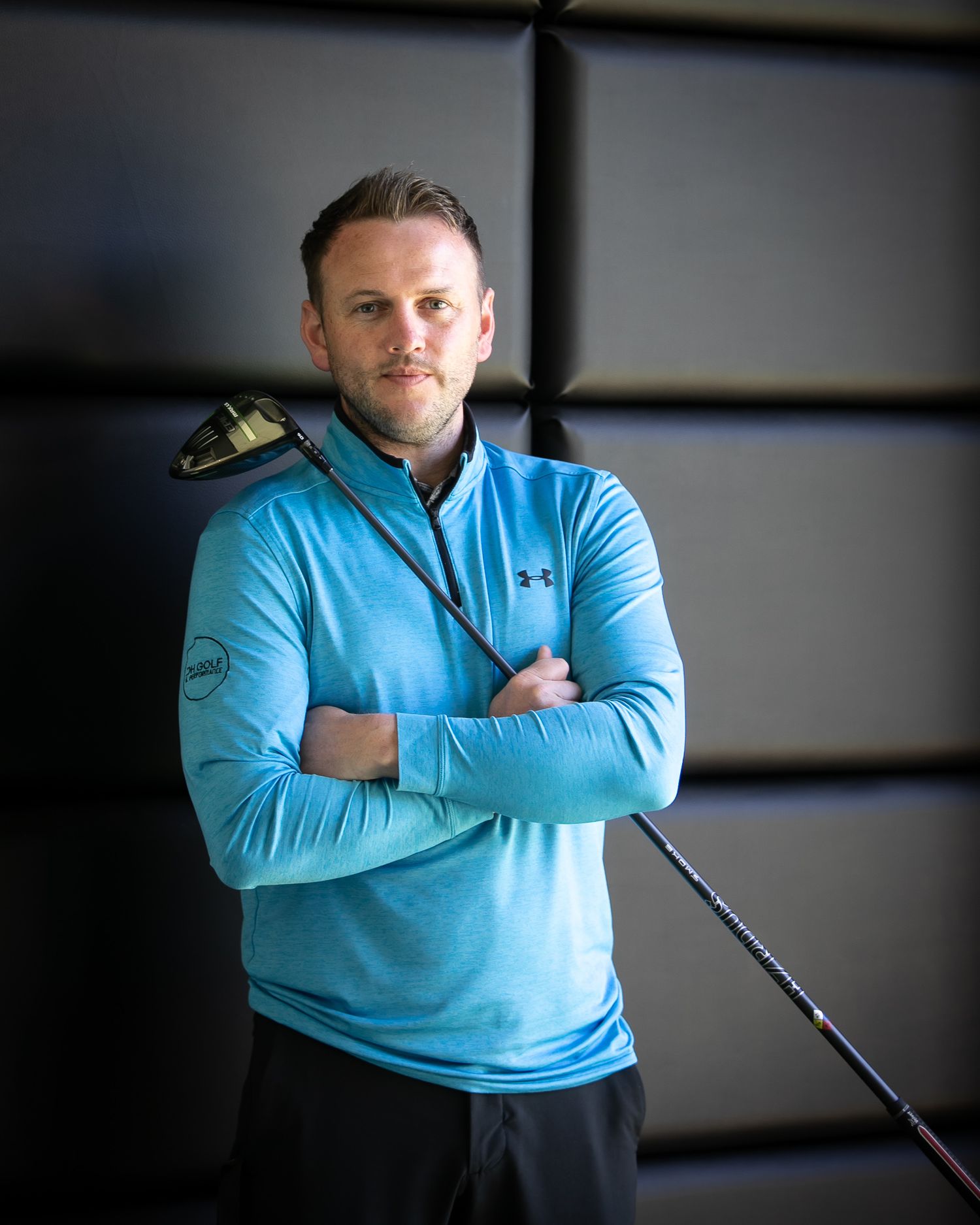 Golfer in light blue Under Armour pullover holds club while posing with crossed arms in front of dark background.