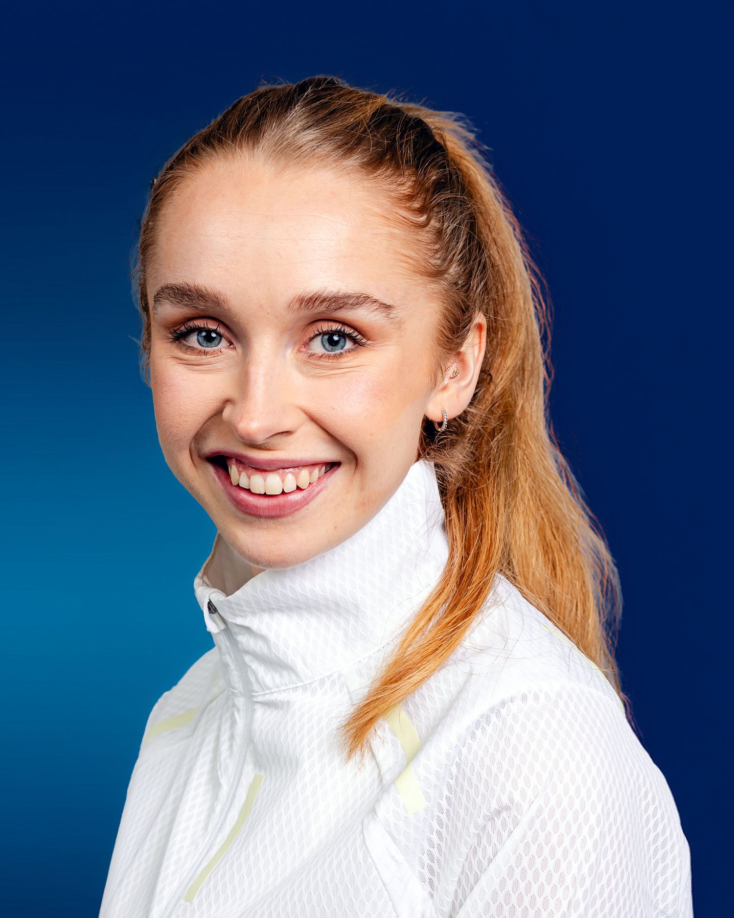 A person in a white turtleneck sweater smiles warmly against a deep blue background.