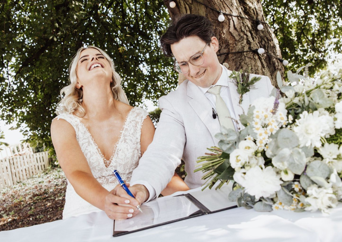 Wedding couple signing marriage documents outdoors with white flowers on table.