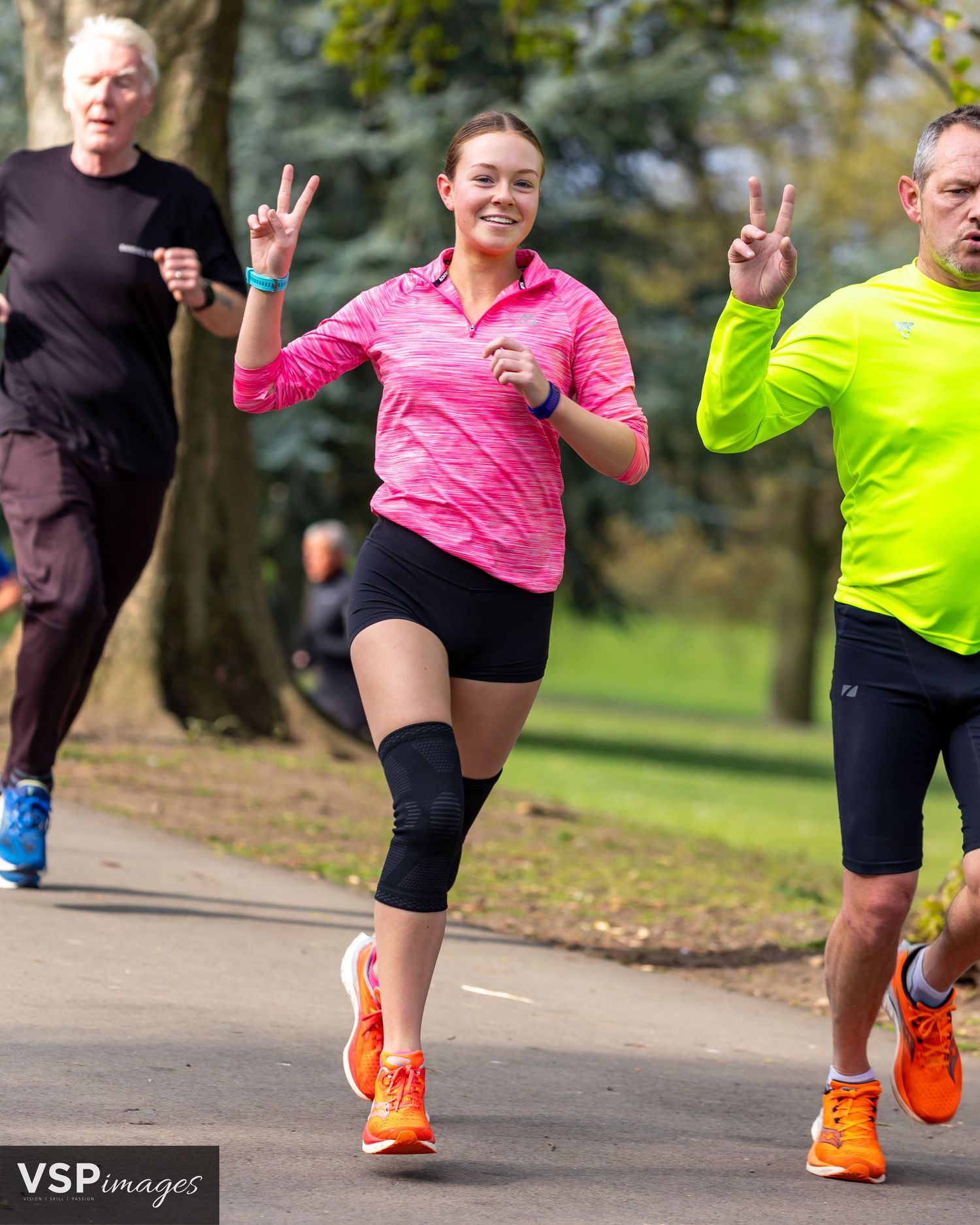 Three runners in bright athletic wear making peace signs while jogging on a park path.