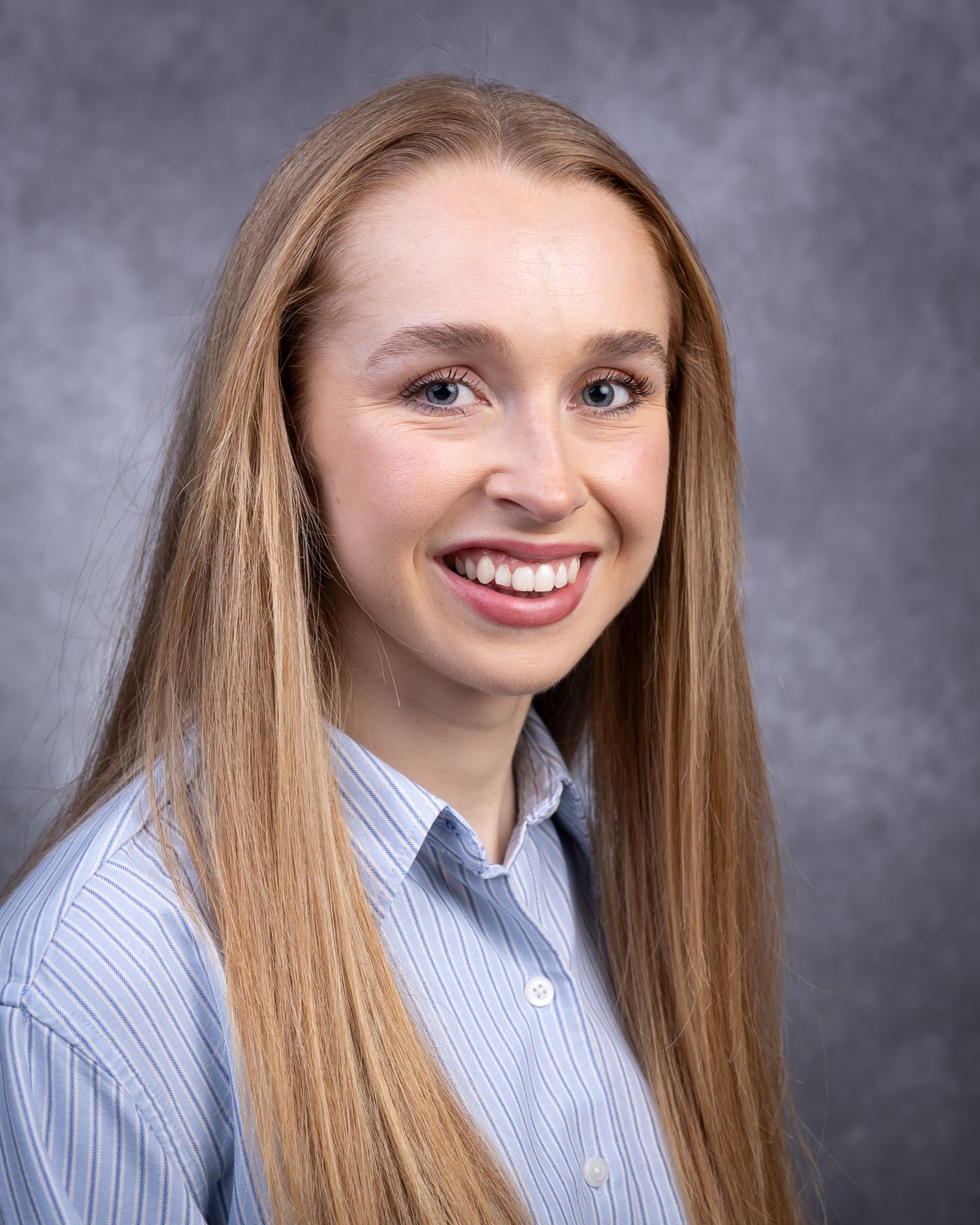 Professional headshot portrait of someone with long blonde hair wearing a light blue button-down shirt against a gray backdrop.