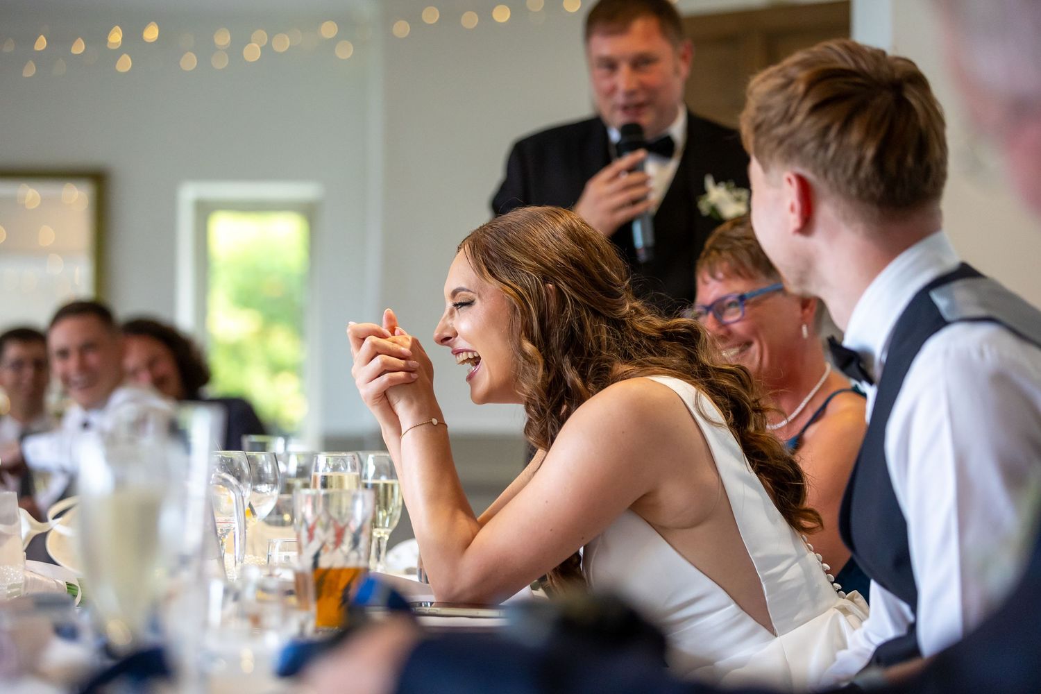 People at a wedding reception enjoying food and drinks at tables decorated with string lights overhead.