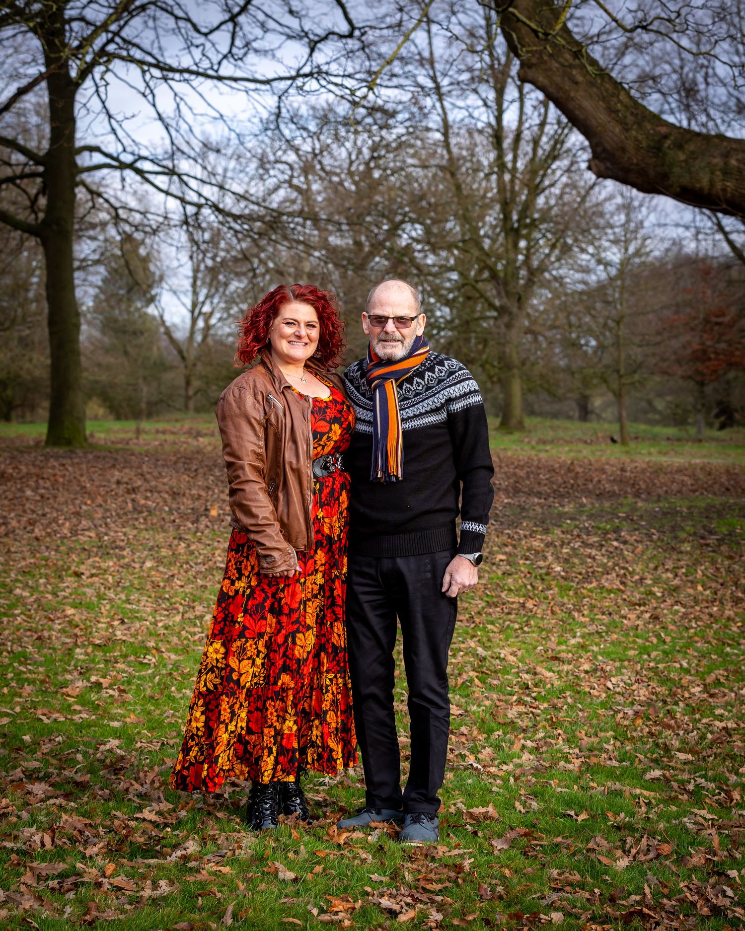 A couple stands together on fallen autumn leaves in a park on an overcast day.