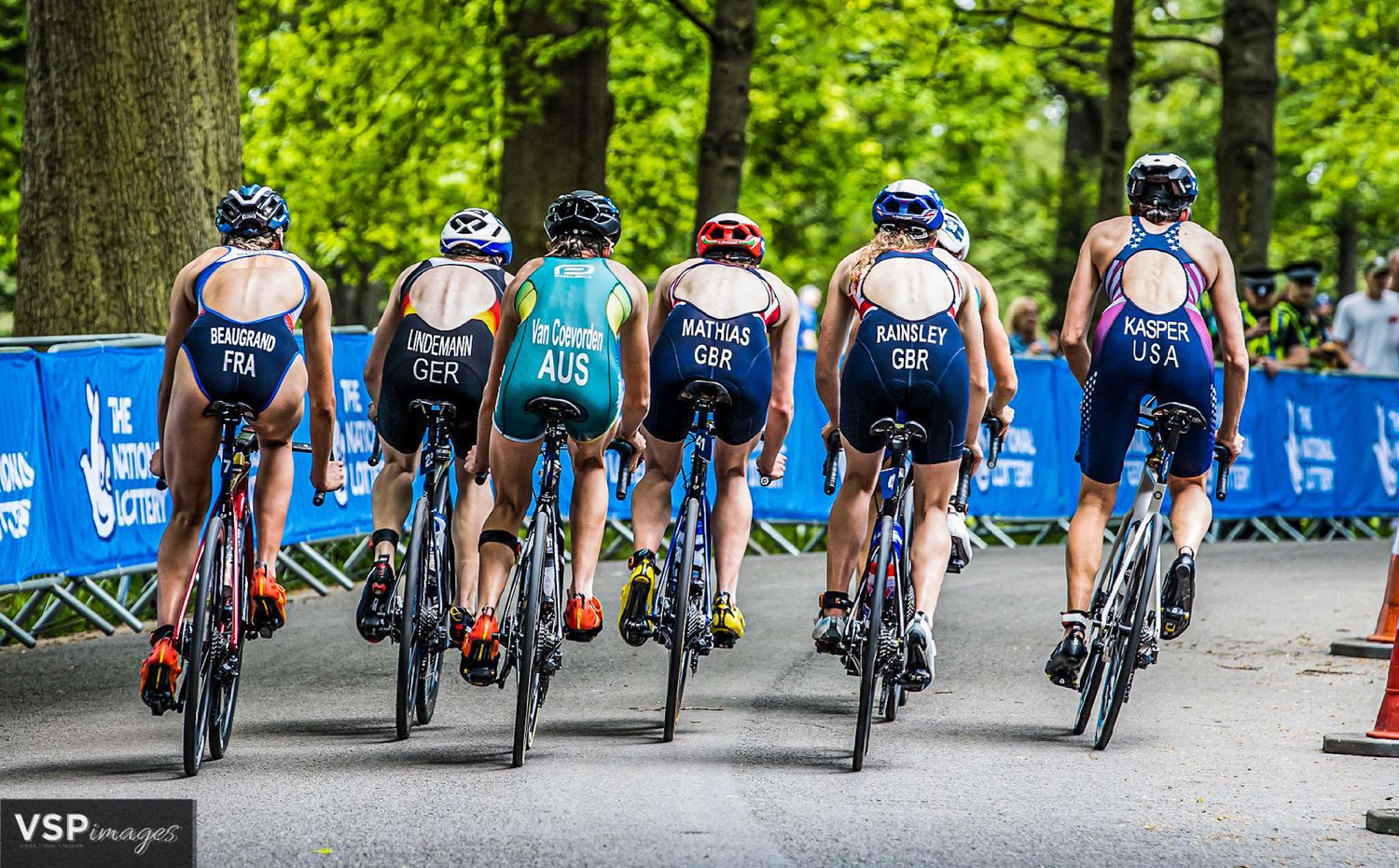 Cyclists in blue uniforms racing on road bikes through a green forested course during a competition event.
