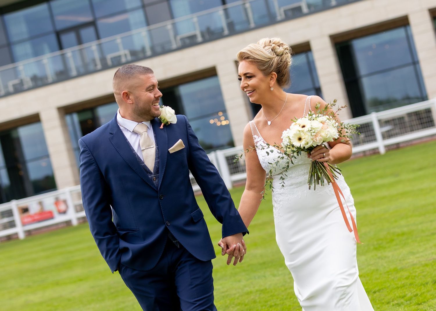 Playful wedding couple having fun on manicured lawn with modern building in background.