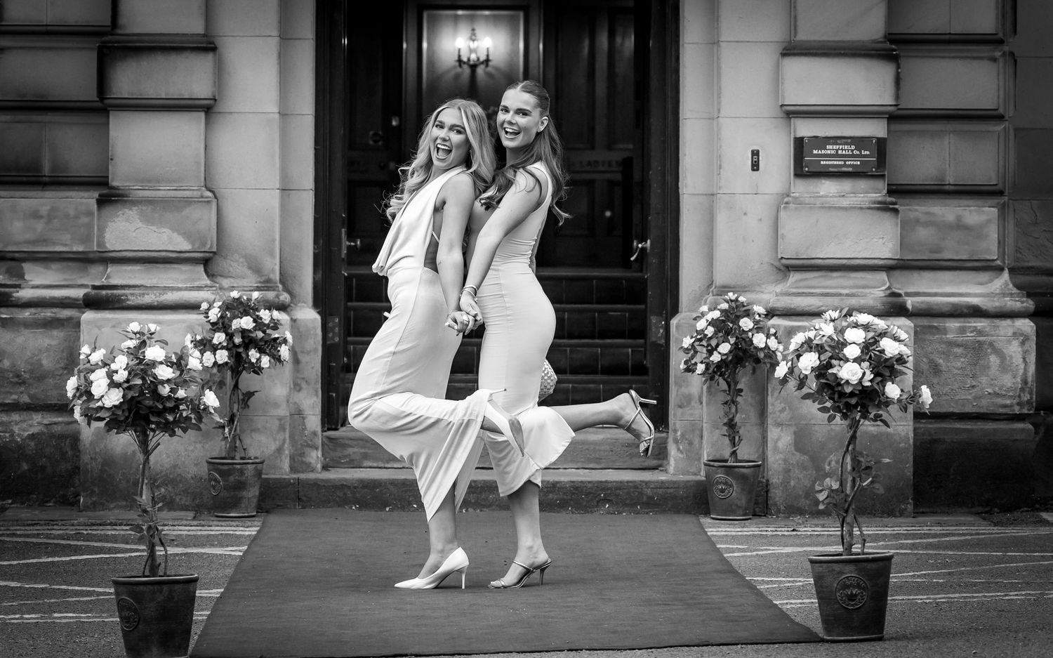 Black and white photo of two dresses posing playfully outside elegant building entrance.