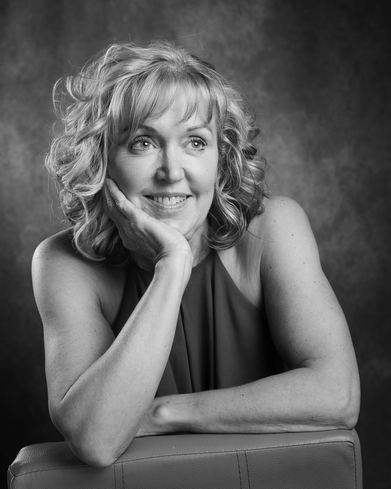 Black and white portrait of a smiling middle-aged blonde woman resting her chin on her hand.