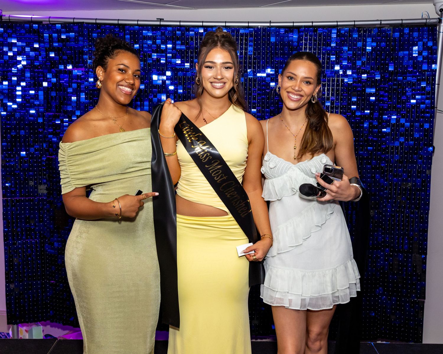 Three young women posing together at a formal event, middle woman wearing a sash, blue sequin backdrop.