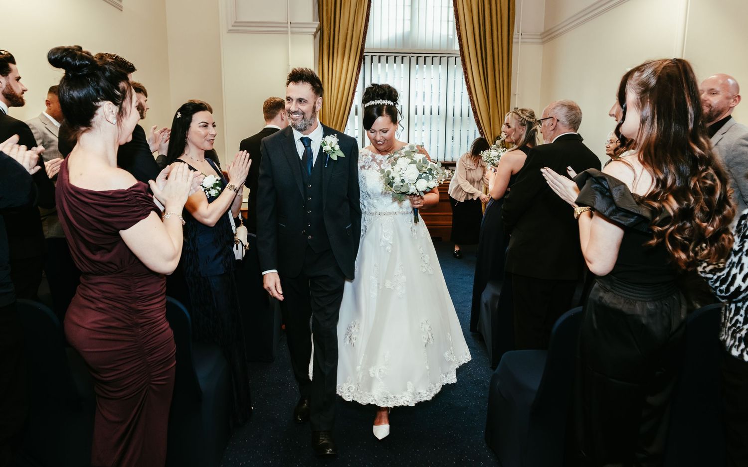 Guests dance and celebrate at an elegant wedding reception with dark curtains and formal attire.