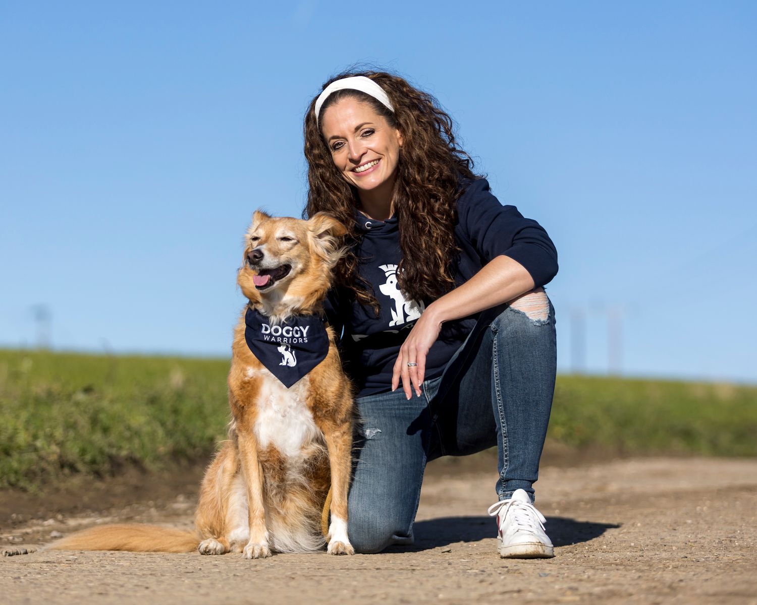Brown mixed breed dog wearing navy bandana sitting outdoors on dirt path.